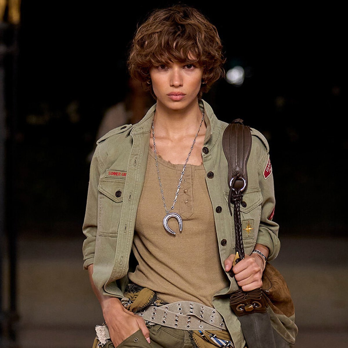Model on a runway wearing an olive military-style jacket over a beige knit top, with layered necklaces and a shoulder bag strapped across.