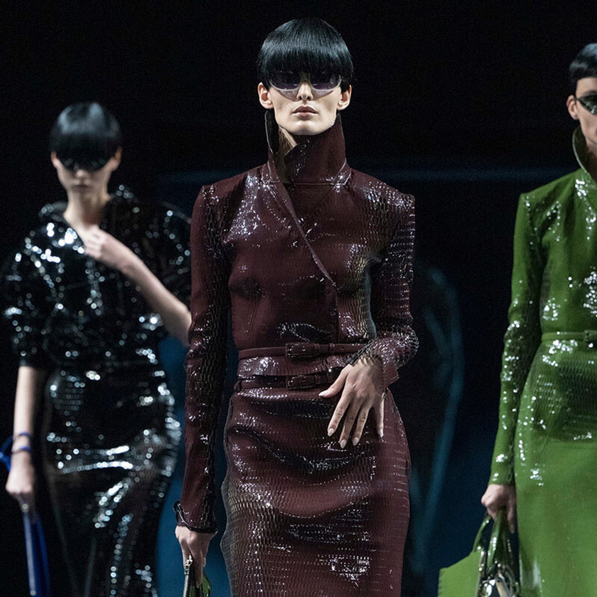 Model on runway wearing a burgundy glossy, high-collar dress with belt, flanked by other models in black and green sequined outfits.