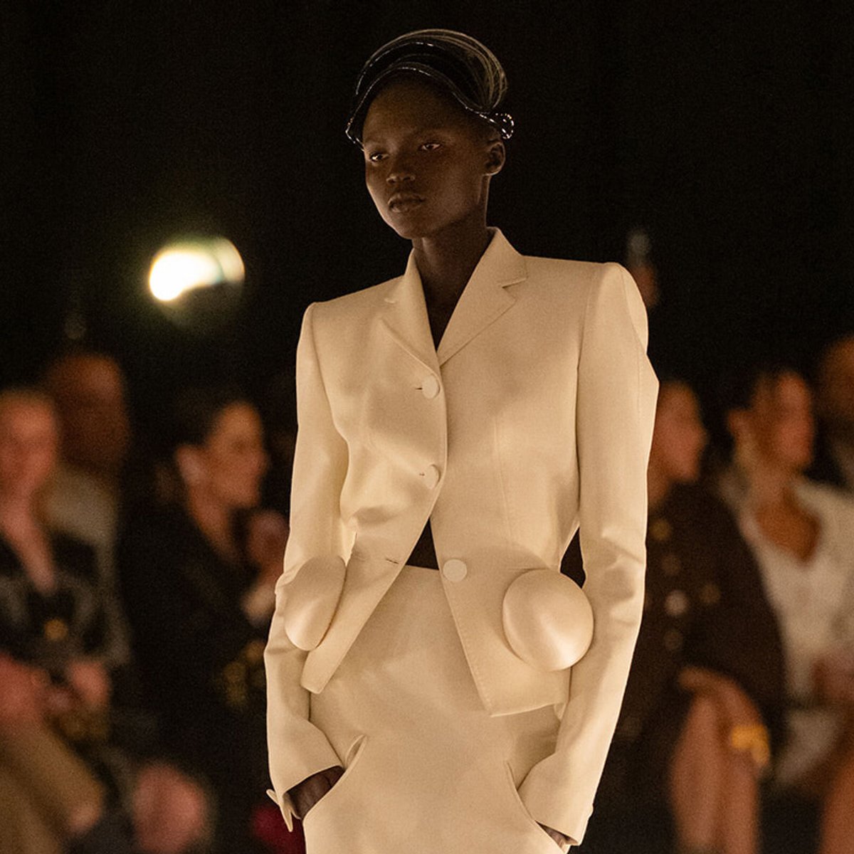Model on a runway wearing a cream tailored suit with large round pocket flaps and a sculptural hat, during a fashion show.