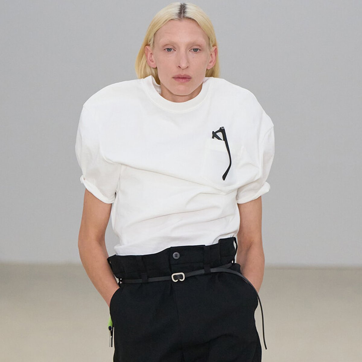 Model wearing an oversized white T-shirt with rolled sleeves and black belted pants, glasses tucked in the chest pocket.