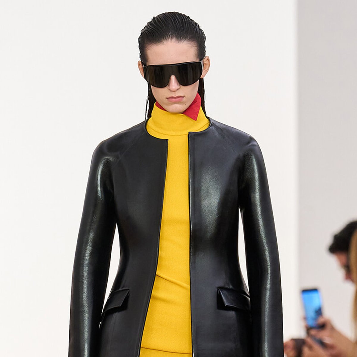 Model on a runway wearing a black leather jacket over a bright yellow outfit with a red collar detail and oversized black sunglasses.