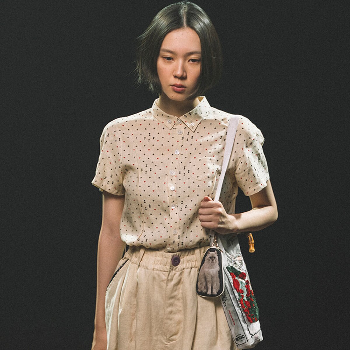 Young woman wearing a beige polka-dot blouse and light beige skirt, carrying a white shoulder bag with a small animal-print pouch against a dark background, in a fashion pose.