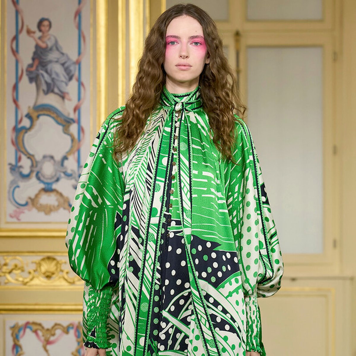 Model walking the runway in a green and white patterned cape-style dress with pink eye makeup, ornate gold-trimmed backdrop behind.