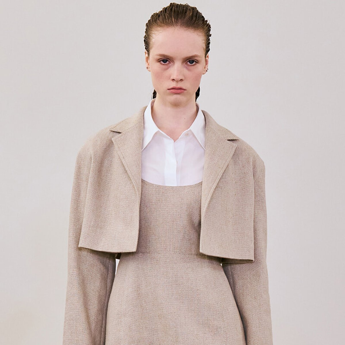 Female model wearing a beige textured coat over a white shirt and matching beige dress, posing with a serious expression against a neutral backdrop.