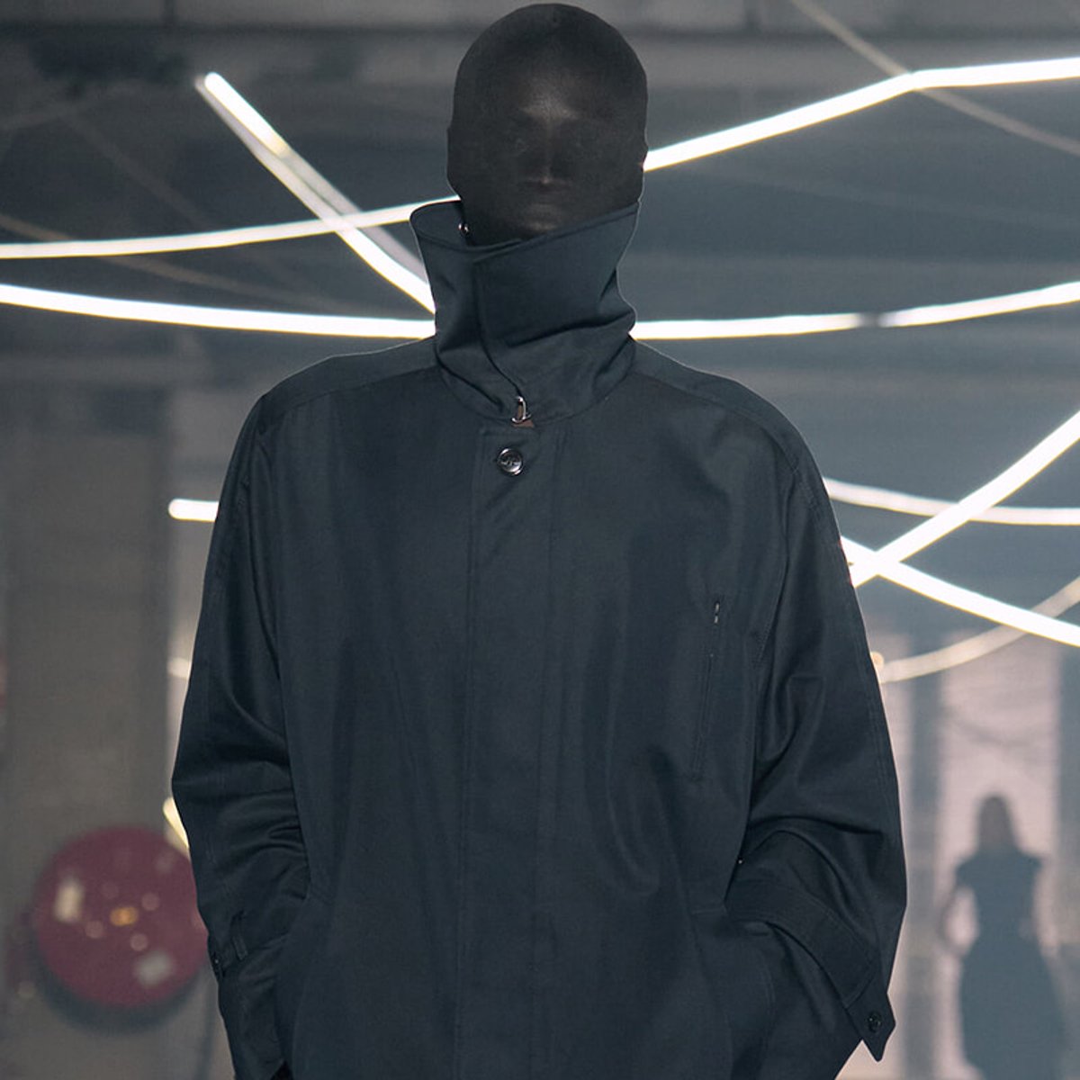 Model wearing a dark high-collar coat in a dim, neon-lit showroom with futuristic lighting behind.