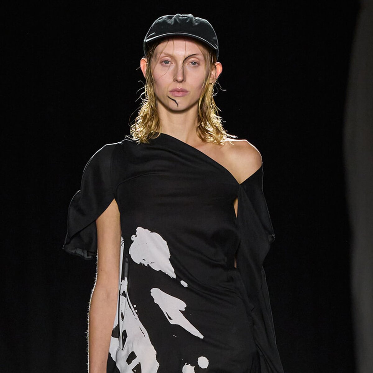 Female model walking a fashion show wearing a black off-shoulder dress with white abstract print and a black cap on a dark runway.