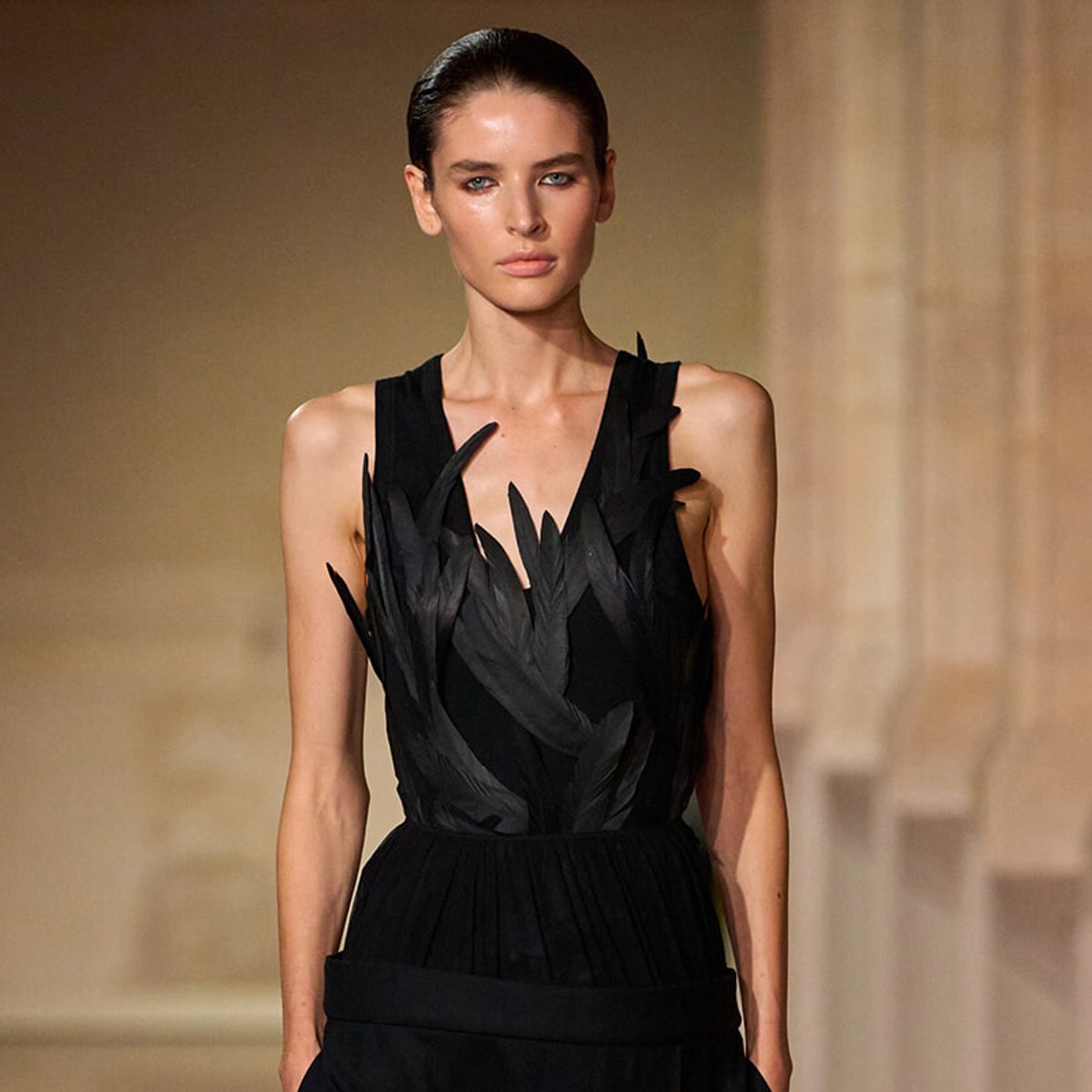 Model on a runway wearing a sleeveless black dress with feather-like appliqués on the bodice.