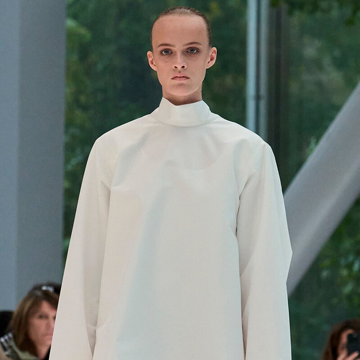 Model walking the runway in a loose, white high-neck blouse or tunic with a minimalist design.