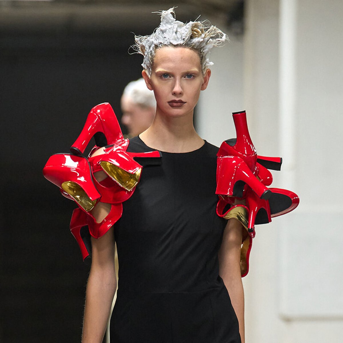 Model wearing a black sheath dress with oversized red glossy sculptural shoulder pieces and silver hair.