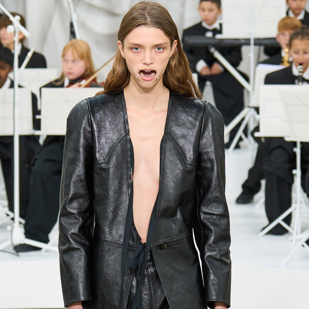 Model wearing a glossy black leather coat with a deep open front walking a fashion show, with musicians in the background.