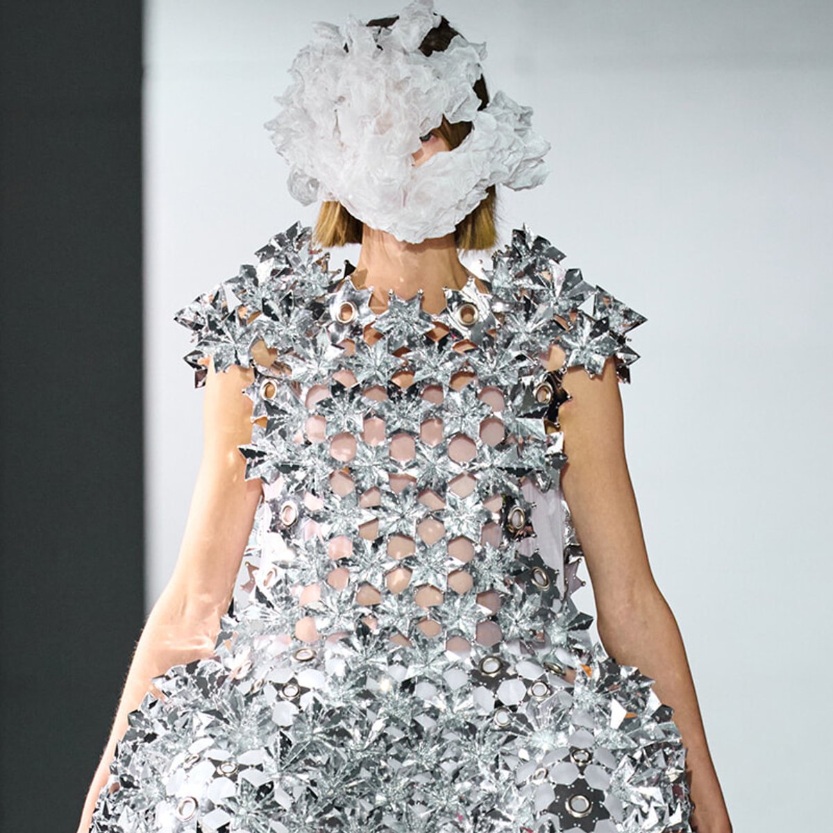 Model wearing a futuristic silver garment made of interlocking star shapes, with a fluffy white cloud-like headpiece obscuring part of the face on a runway.