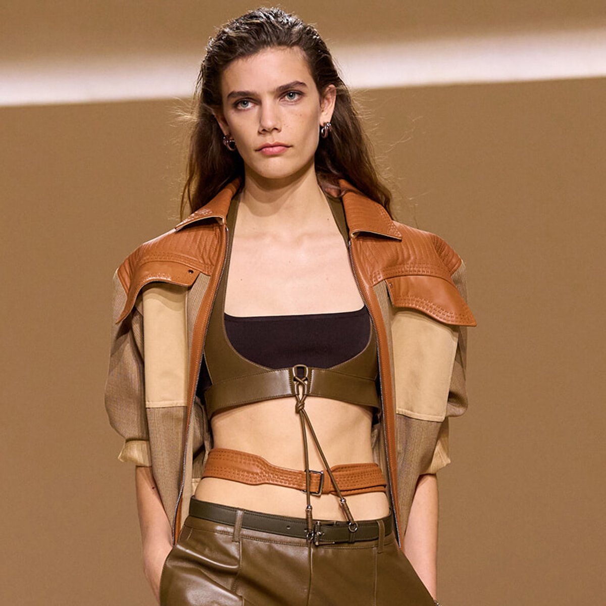 Female model walks the runway in a brown leather cropped harness top, olive-green bralette, and matching high-waisted pants, with a tan leather jacket draped over shoulders.