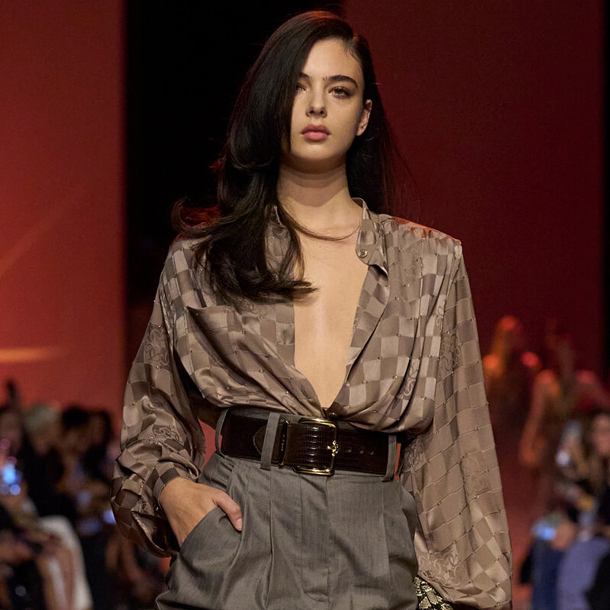 Runway model wearing a taupe satin checkered blouse tucked into gray high-waisted pants with a wide belt.