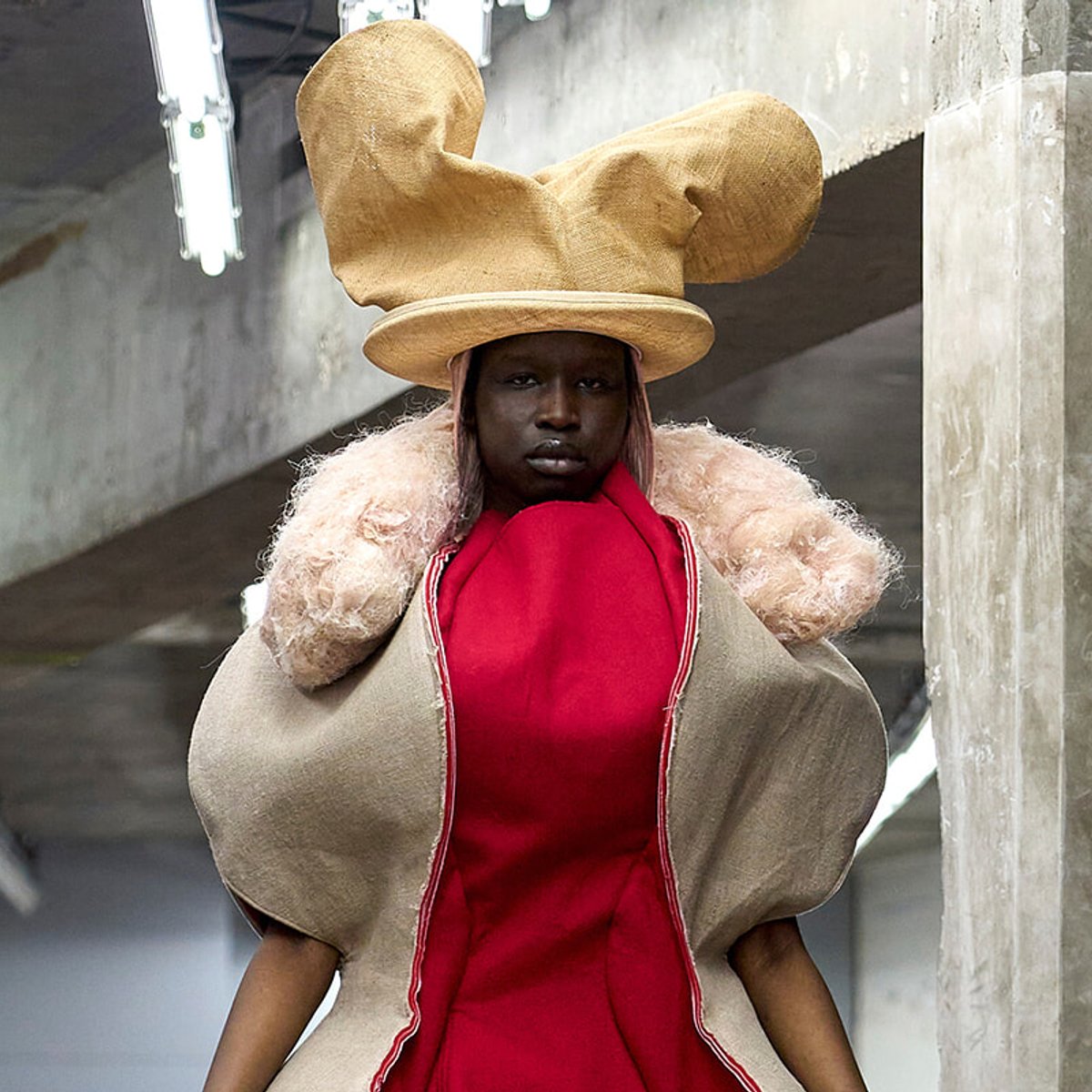 Person wearing a large sculptural beige hat, red dress, and beige coat in an industrial setting.