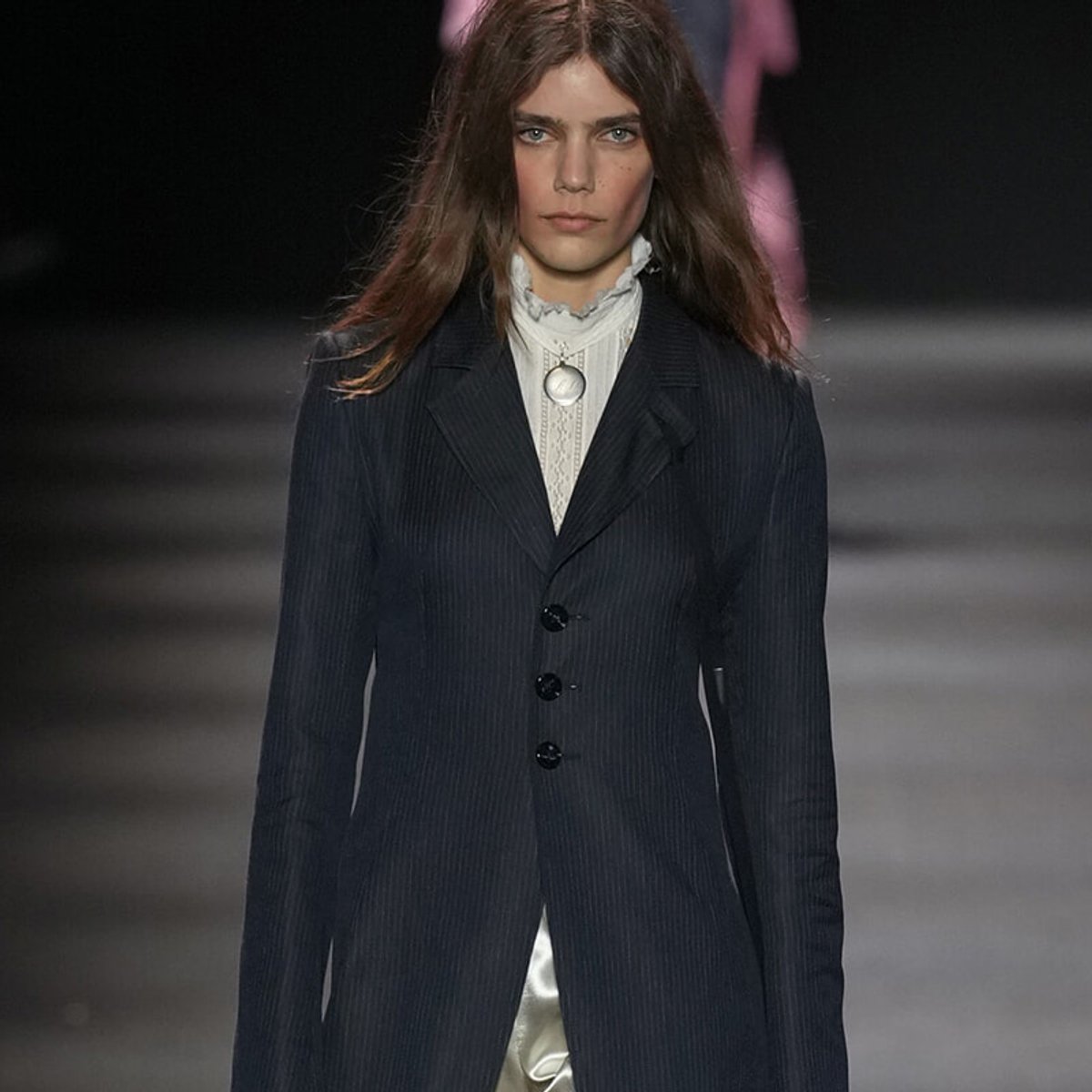 Female model on a runway wearing a dark pinstripe blazer over a white ruffled high-neck blouse with a circular pendant necklace on a chain.