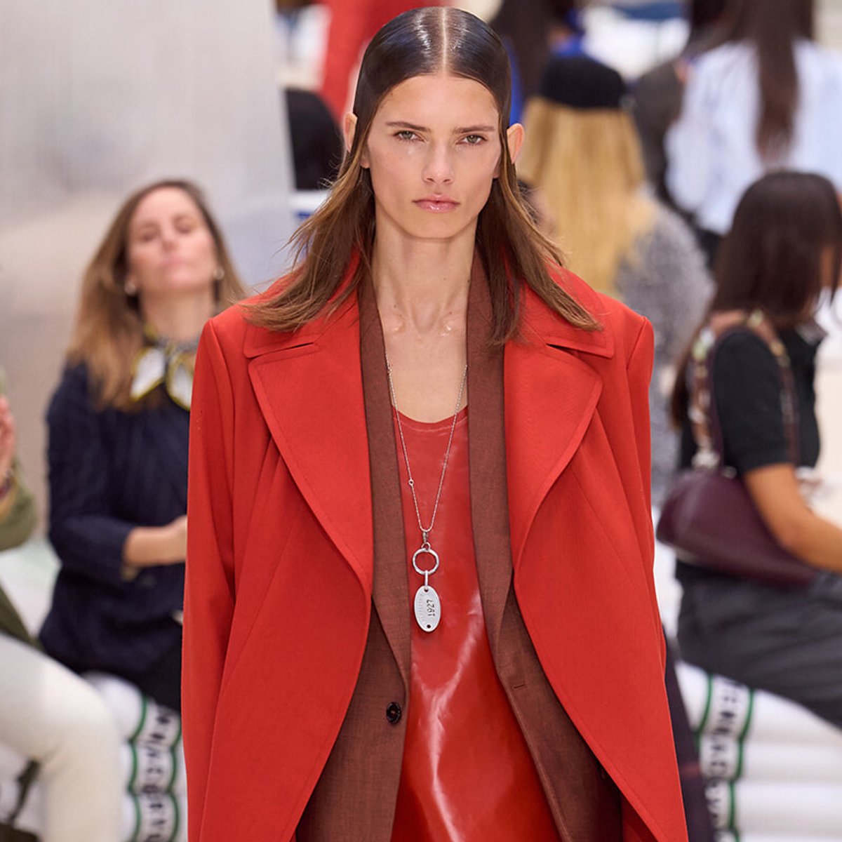 Model walking a runway in a bold red coat over a brown suit, silver pendant necklace visible at chest level, blurred audience in background.