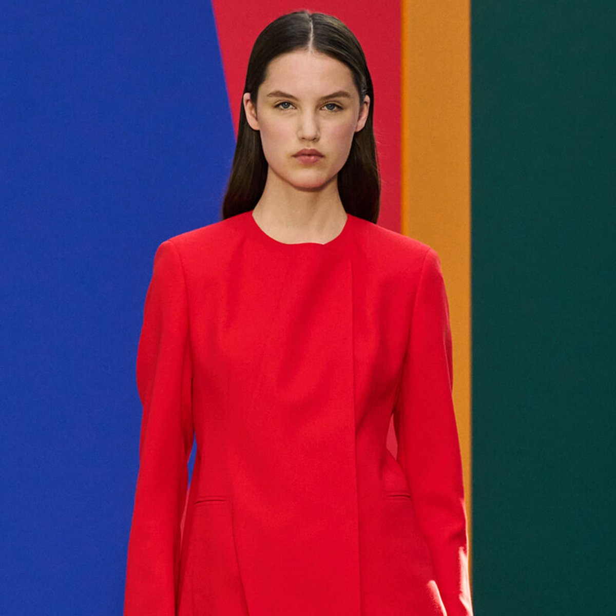 Model in a bright red dress standing against a colorful striped backdrop (blue, red, orange, green) creating a bold fashion look.