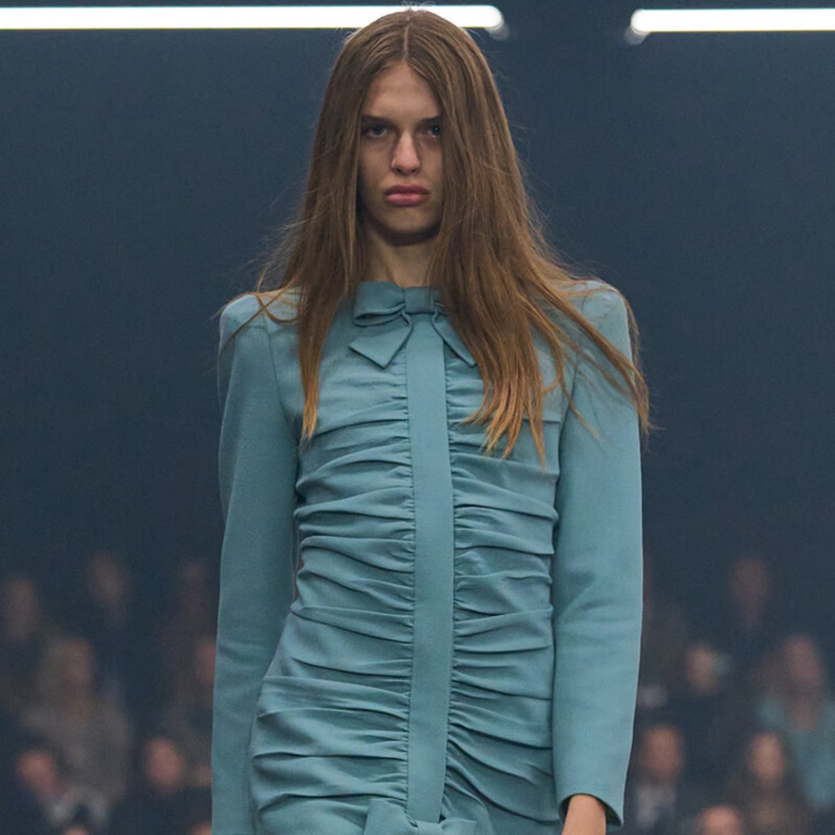 Model wearing a light teal ruched dress with a bow at the neck on a runway (catwalk).