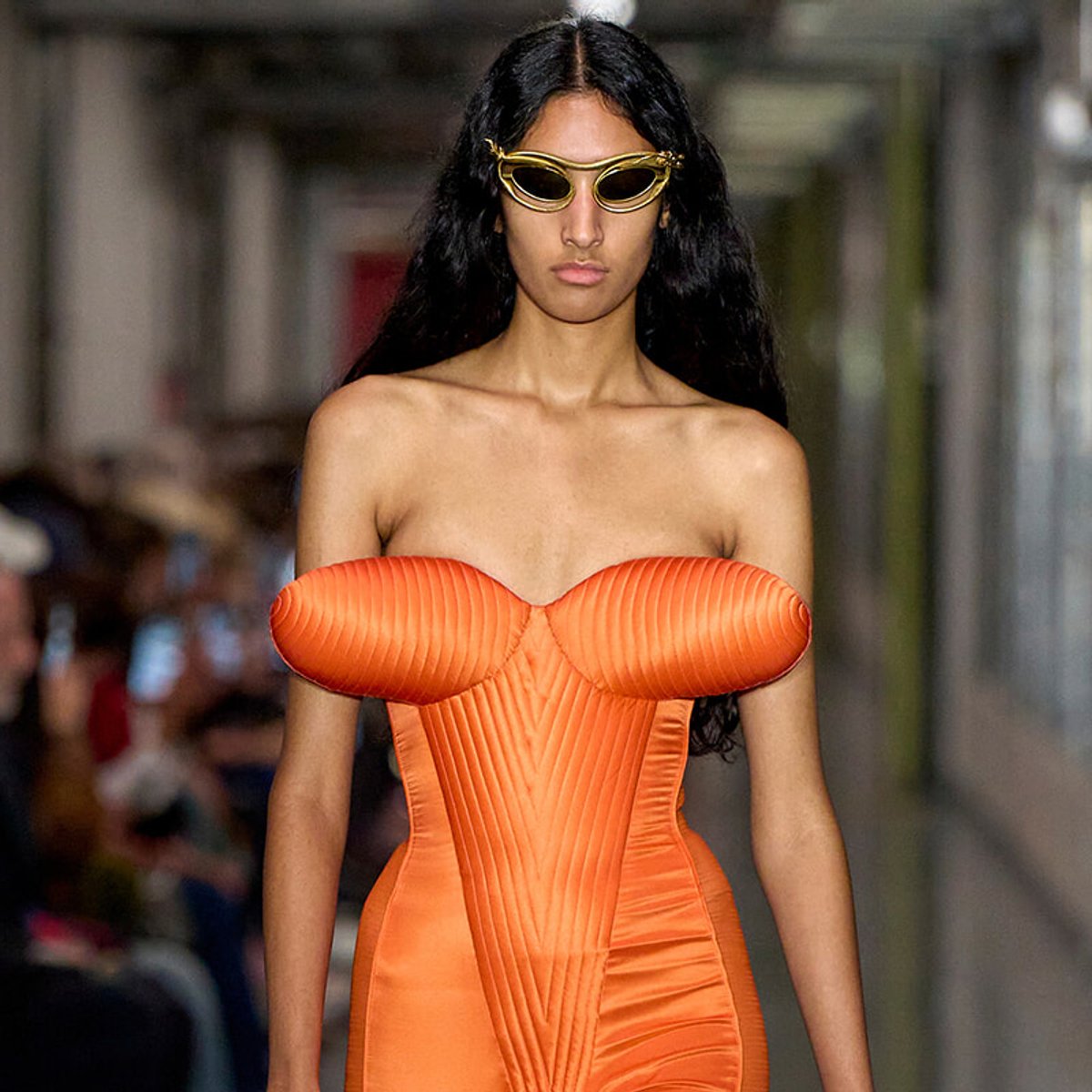 Female model walks a runway in a bright orange strapless corset dress with gold sunglasses.