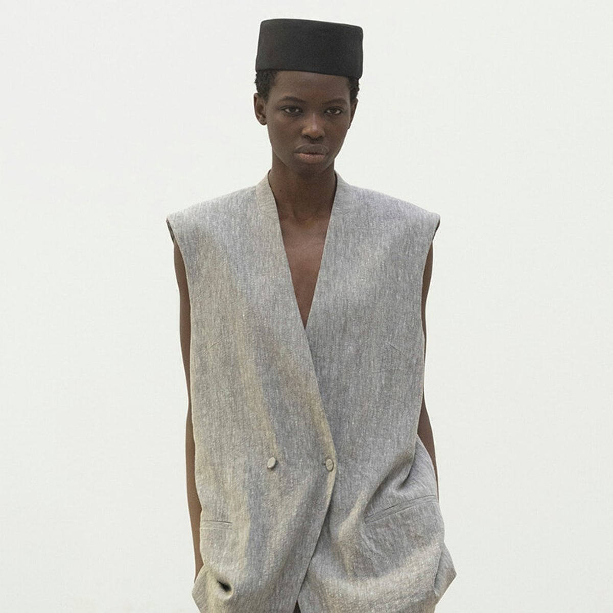Model wearing a light gray sleeveless blazer with a deep V, paired with a black fez-style hat, standing against a plain background.