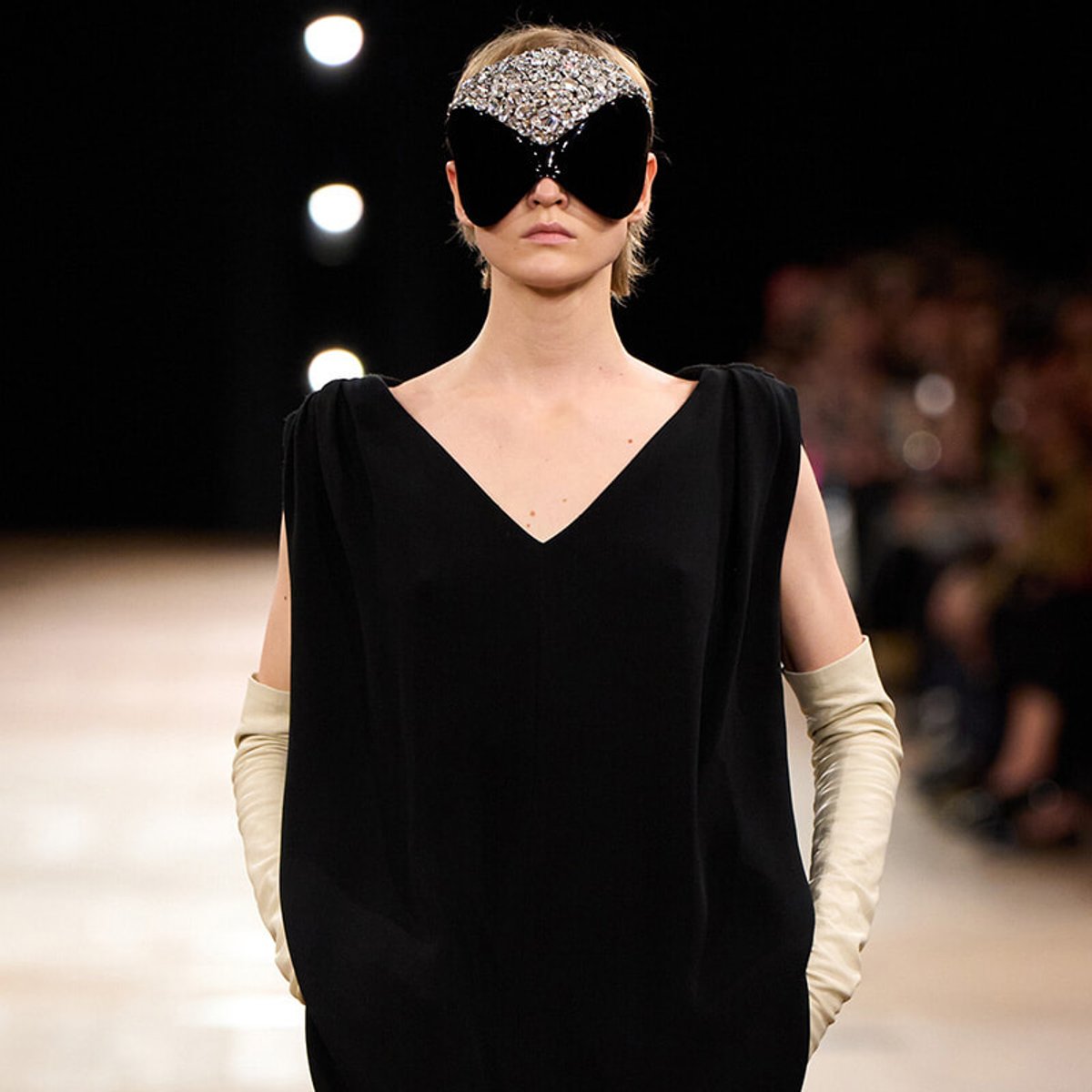 Runway model in a black V-neck dress with long cream gloves and a jeweled, oversized eye mask/headpiece.