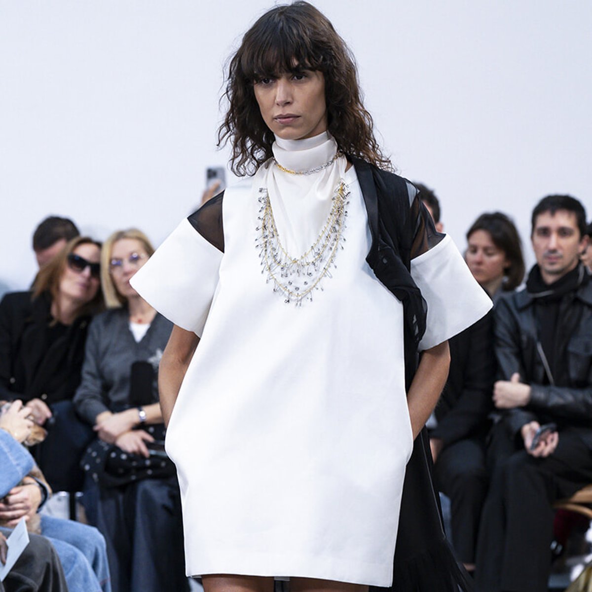 Model on a runway wearing a white loose dress with sheer black sleeves and layered jewelry, audience watching in the background.