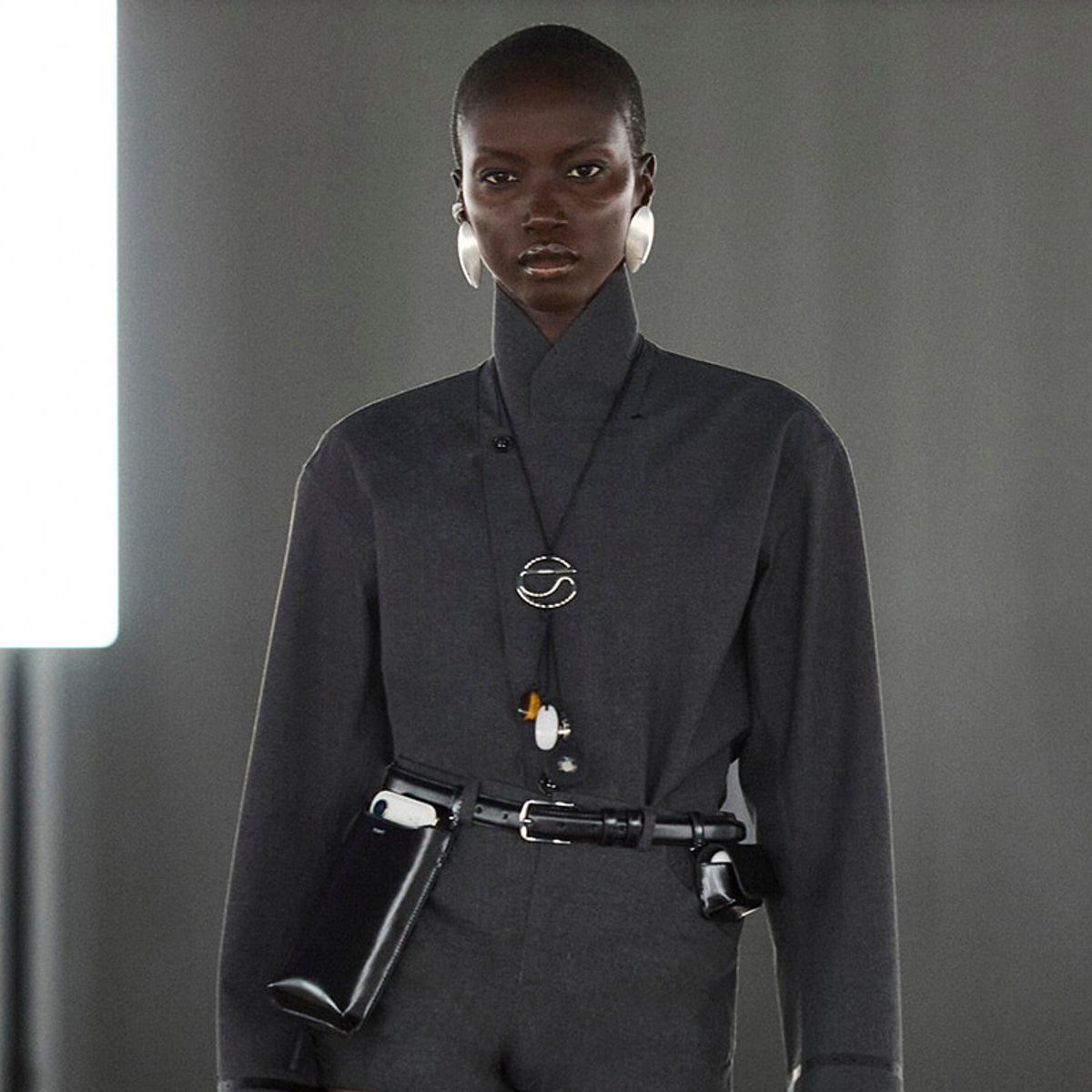 Model in a dark gray high-collar outfit with a black belt, white circular earrings, and a circular pendant necklace in a studio setting.