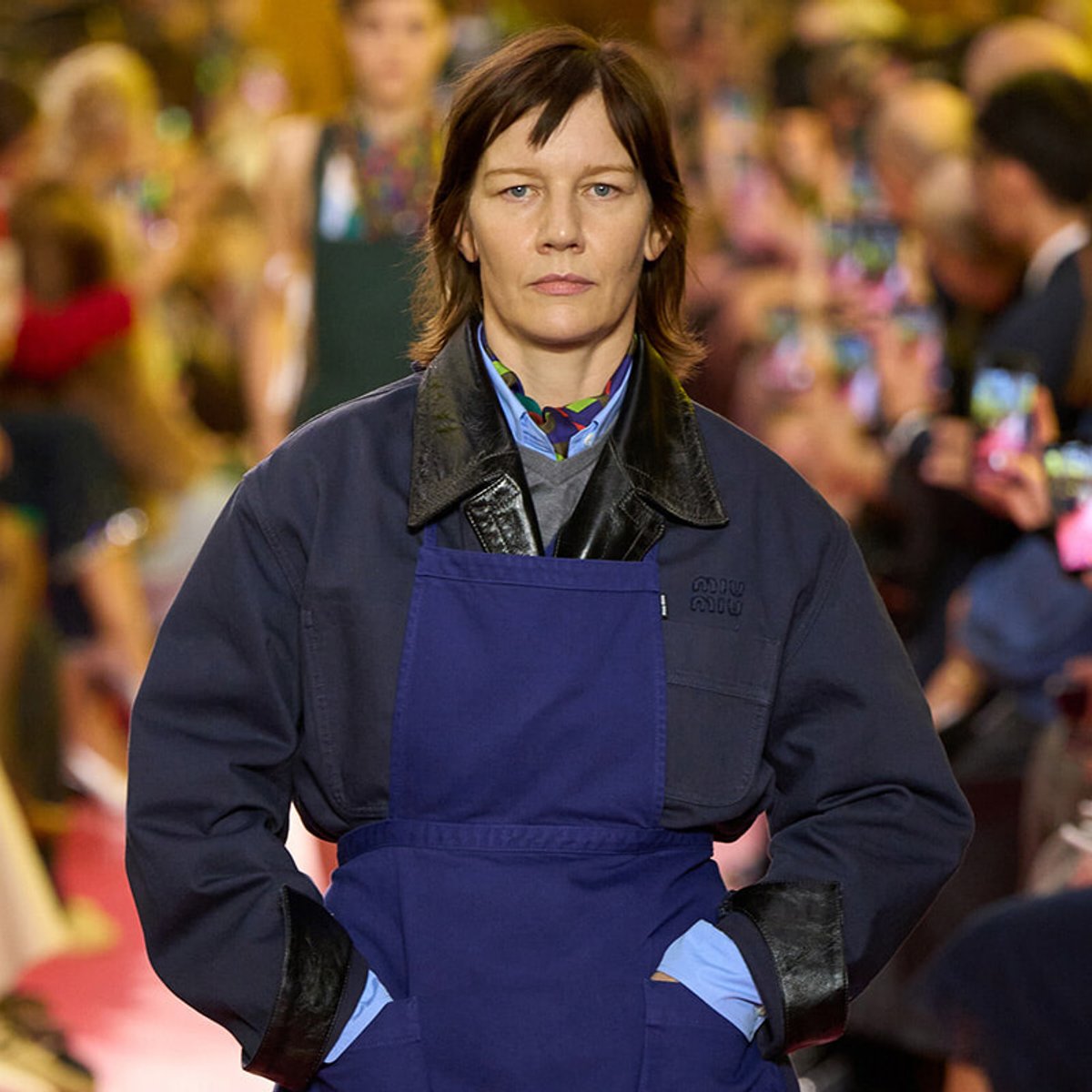 Female model wearing a navy coat with a blue apron, walking on a busy runway while photographers watch in the background.