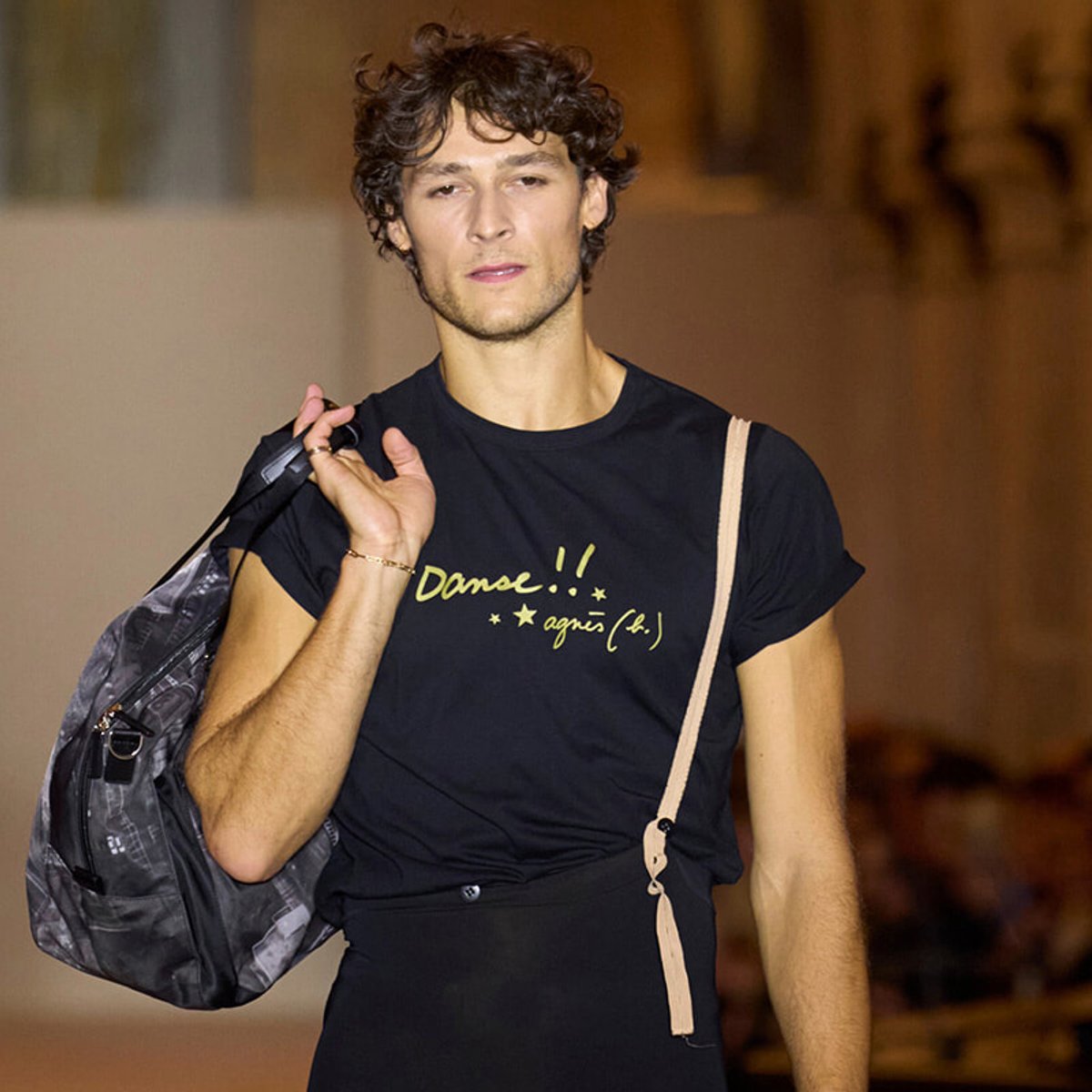 Male model walking a runway with a gray backpack over his shoulder, wearing a black t‑shirt that reads 'Danse!!' in yellow letters.