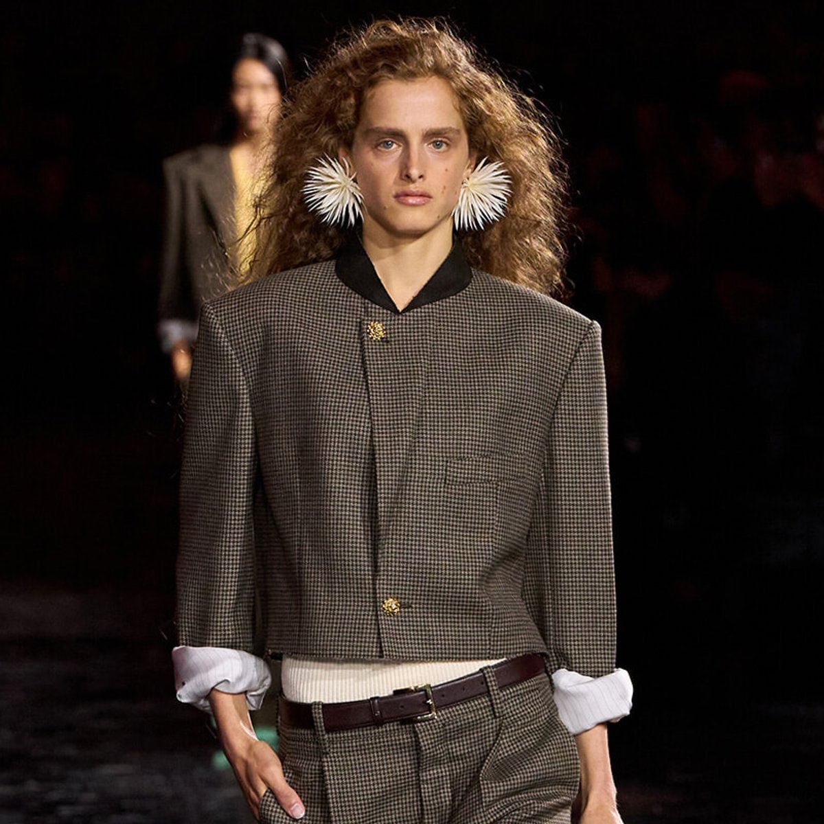 Model walks a fashion runway wearing a brown checkered suit with rolled cuffs and a white knit underlayer, accessorized with large white spiky earrings and a gold brooch