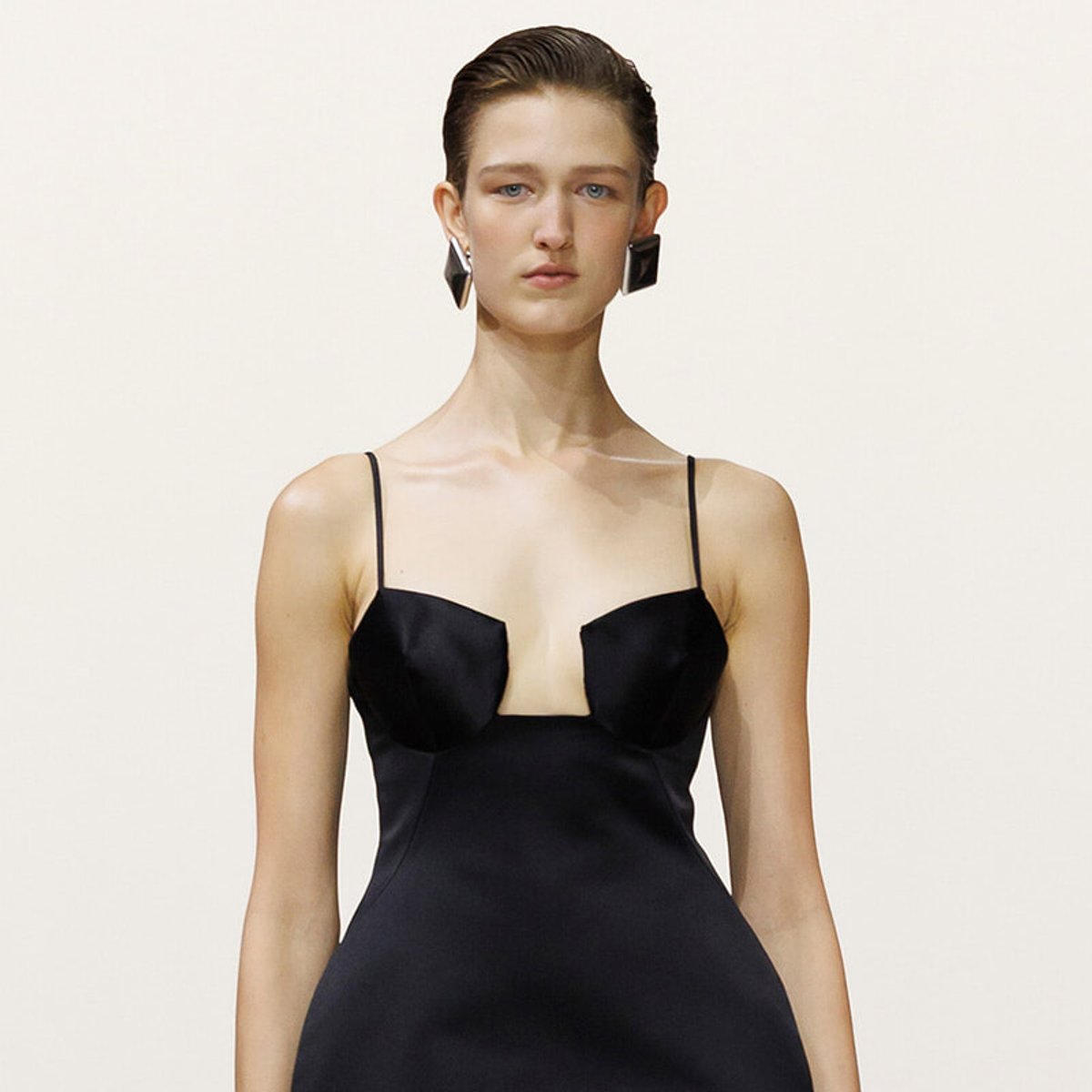 Model on a runway wearing a black satin slip dress with thin straps and a square neckline, accessorized with geometric earrings.