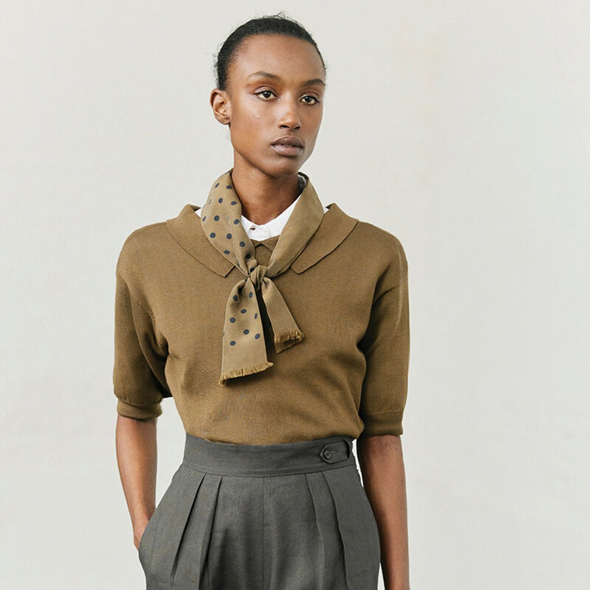 Model wearing a camel-colored knit top with a polka-dot scarf tied at the neck and a gray pleated high-waisted skirt.