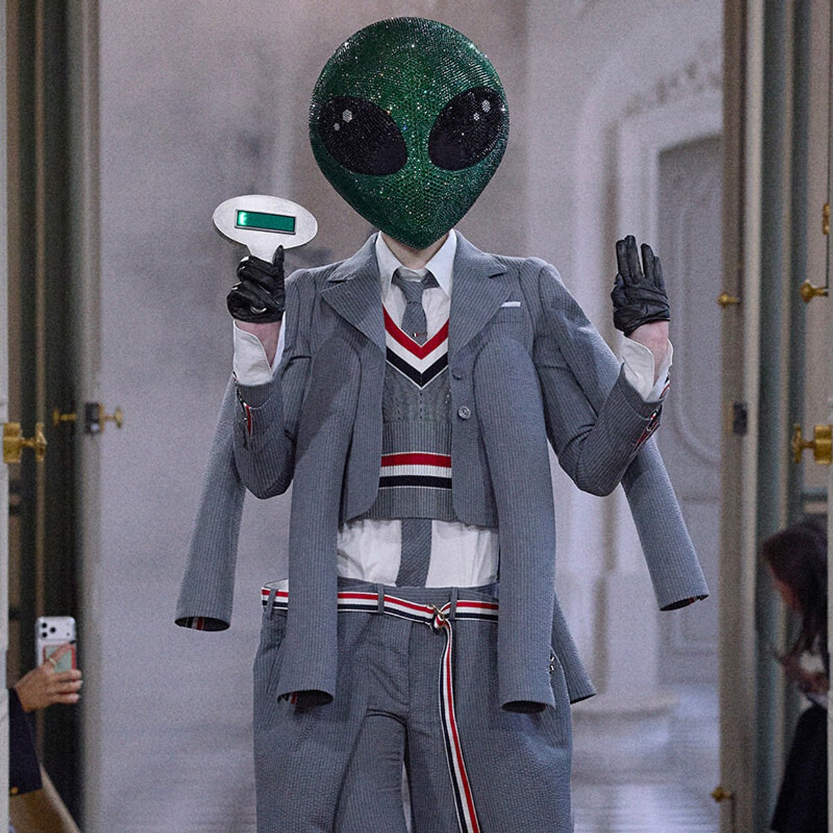 Person wearing a sparkly green alien mask with a gray suit, posing with a peace sign in a hallway.
