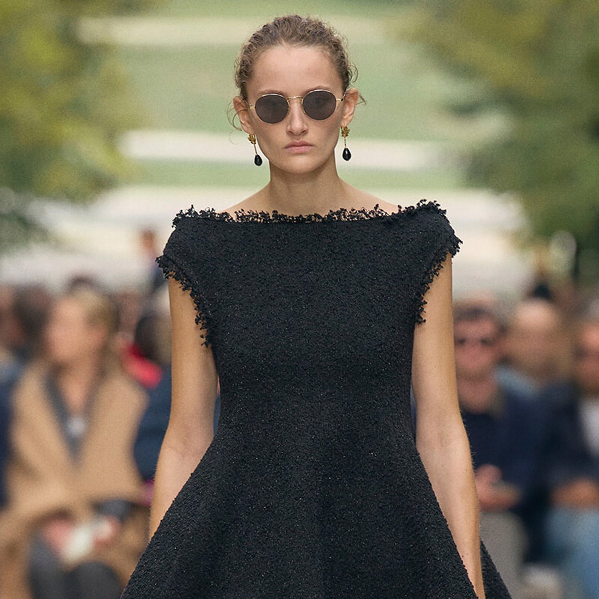 Model walking the runway in a black textured off-the-shoulder gown with round sunglasses and drop earrings.