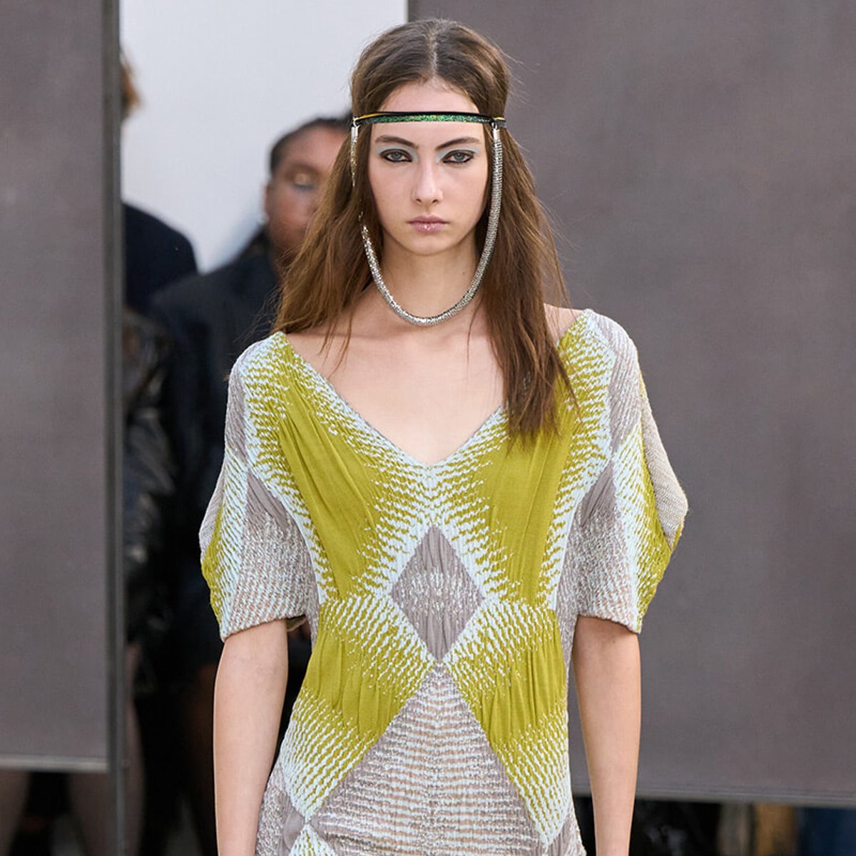 Model on a runway wearing a lime-green and beige geometric knit dress with a green headband and silver necklace.
