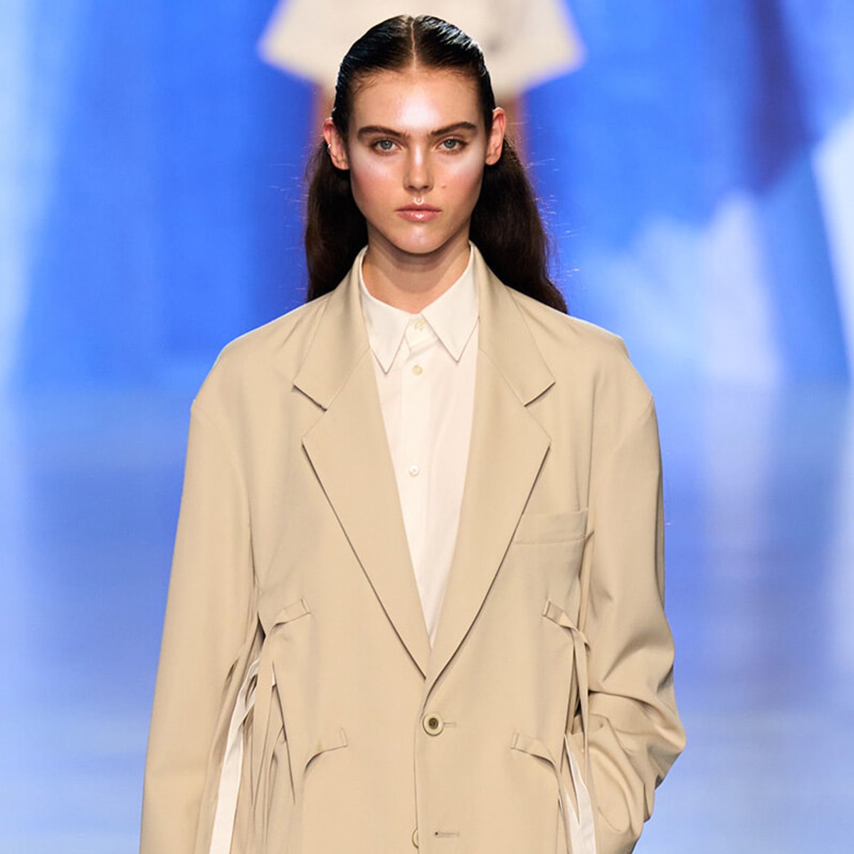 Female model walking a fashion runway in a cream oversized blazer over a white button-down shirt, against a blue backdrop.