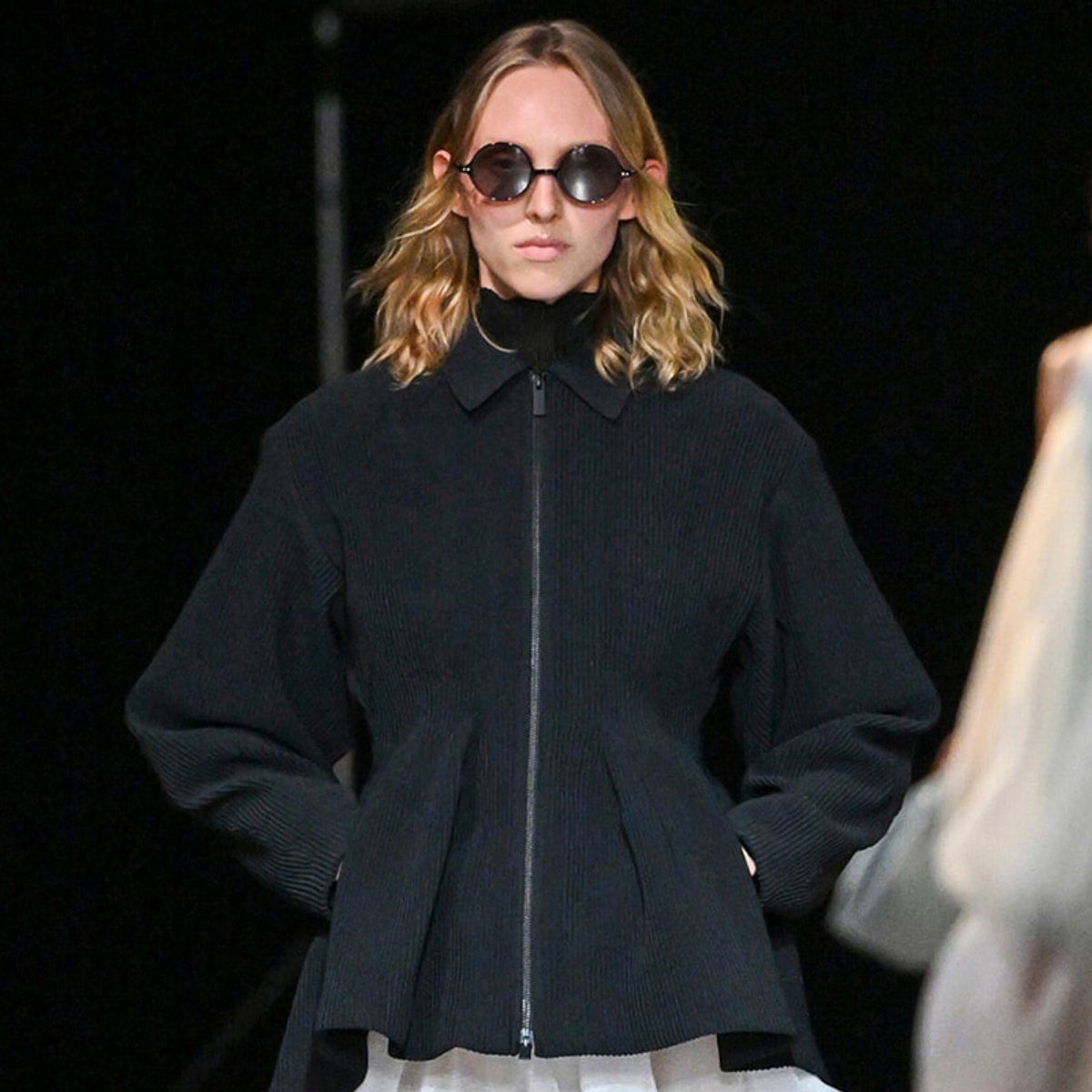 Model on a runway wearing an oversized black zip-front jacket and round sunglasses; hands in pockets, dark background.