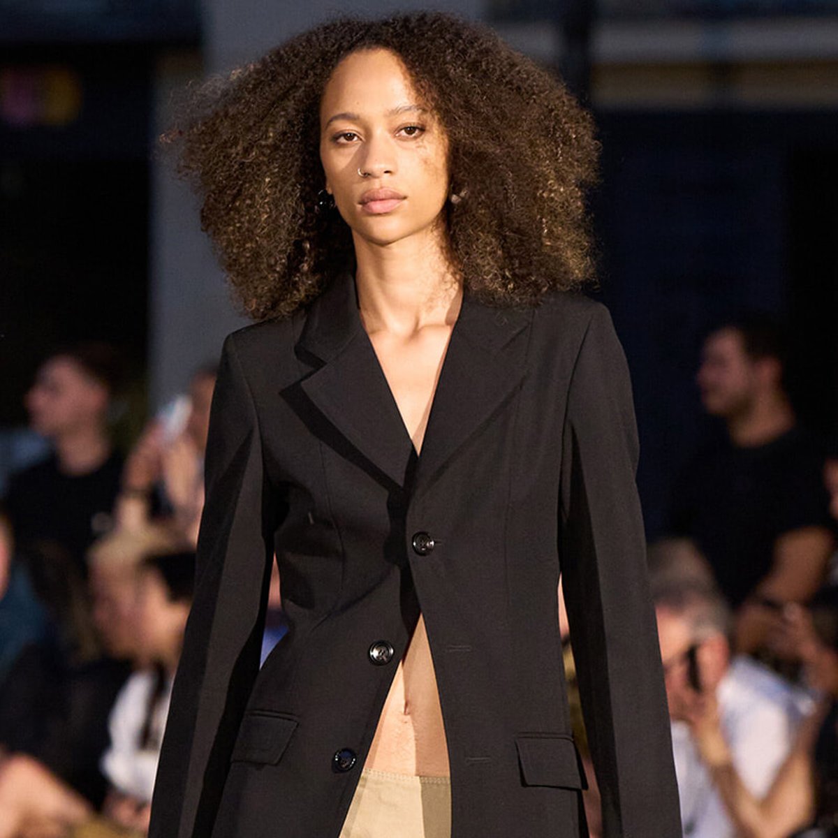 Model walking the runway in a black blazer, audience blurred in the background.