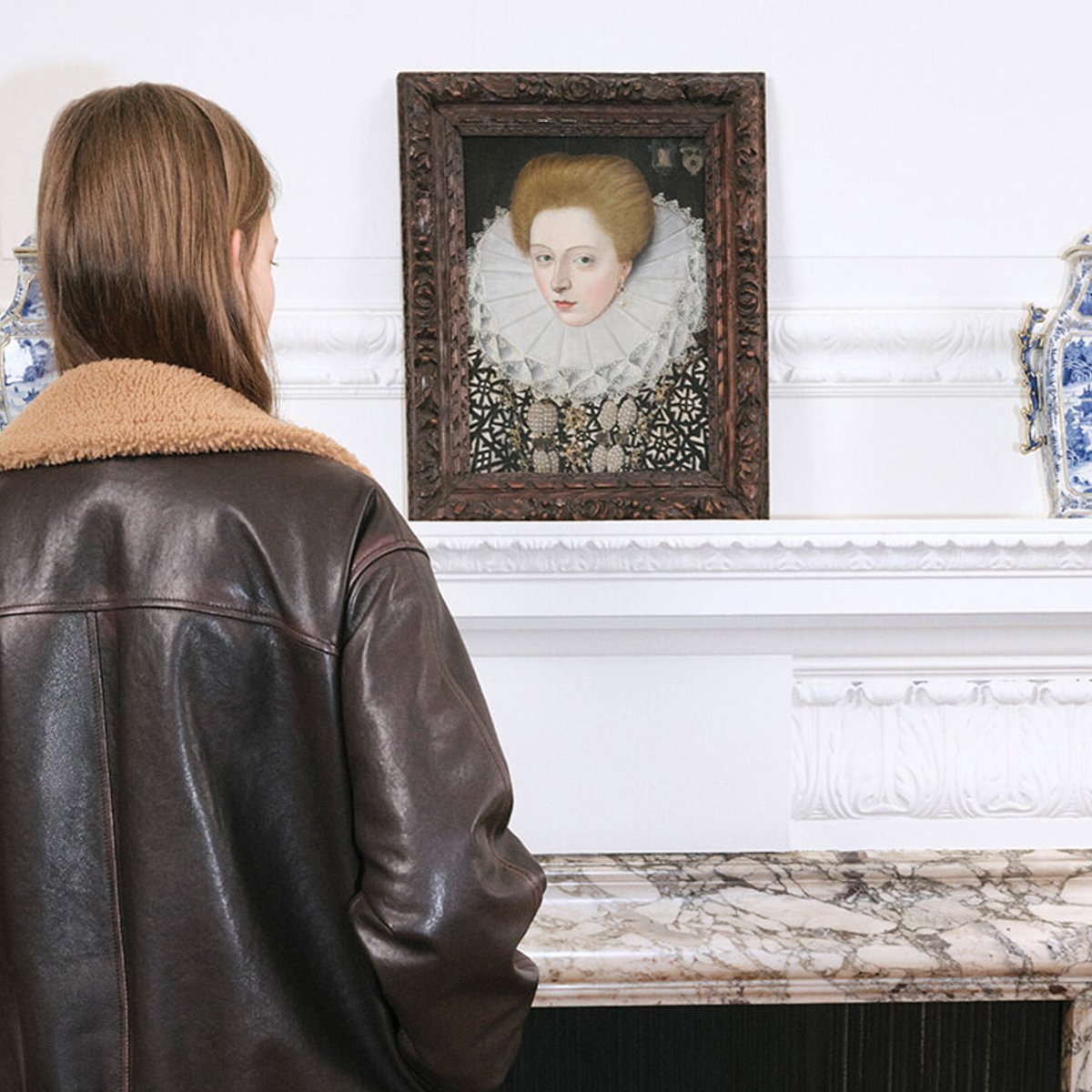 Back view of a person in a brown leather jacket looking at a framed portrait of a woman with a lace collar on a white wall mantel.