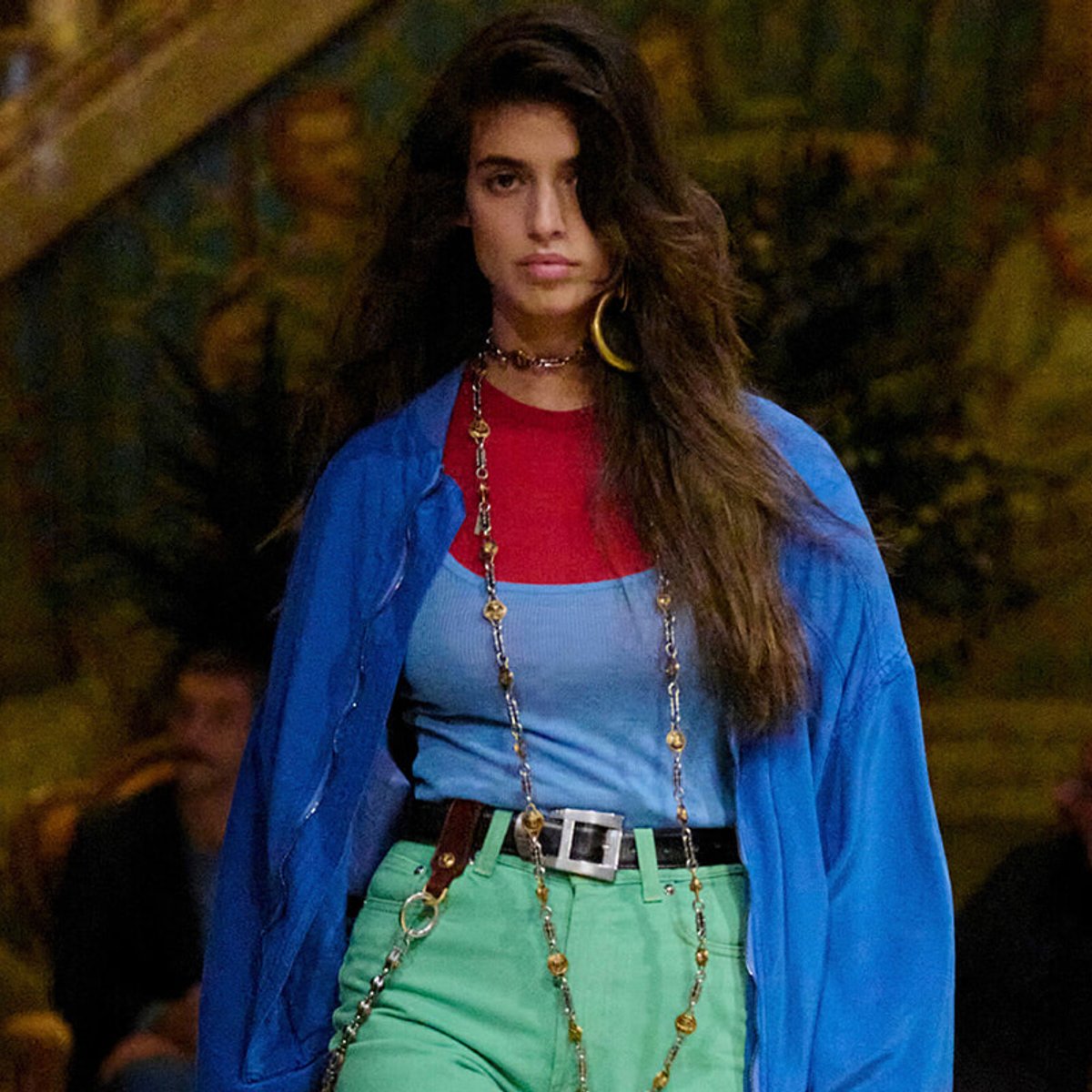 Female model walking runway in a bright blue jacket over a light blue top and red underlayer, paired with green pants and a metallic belt with long beaded chains.