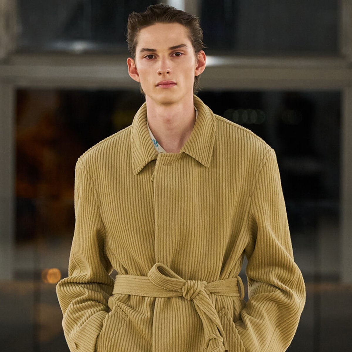 Male model wearing a tan corduroy belted coat with a wide collar, hands in pockets, neutral expression on a fashion runway backdrop.