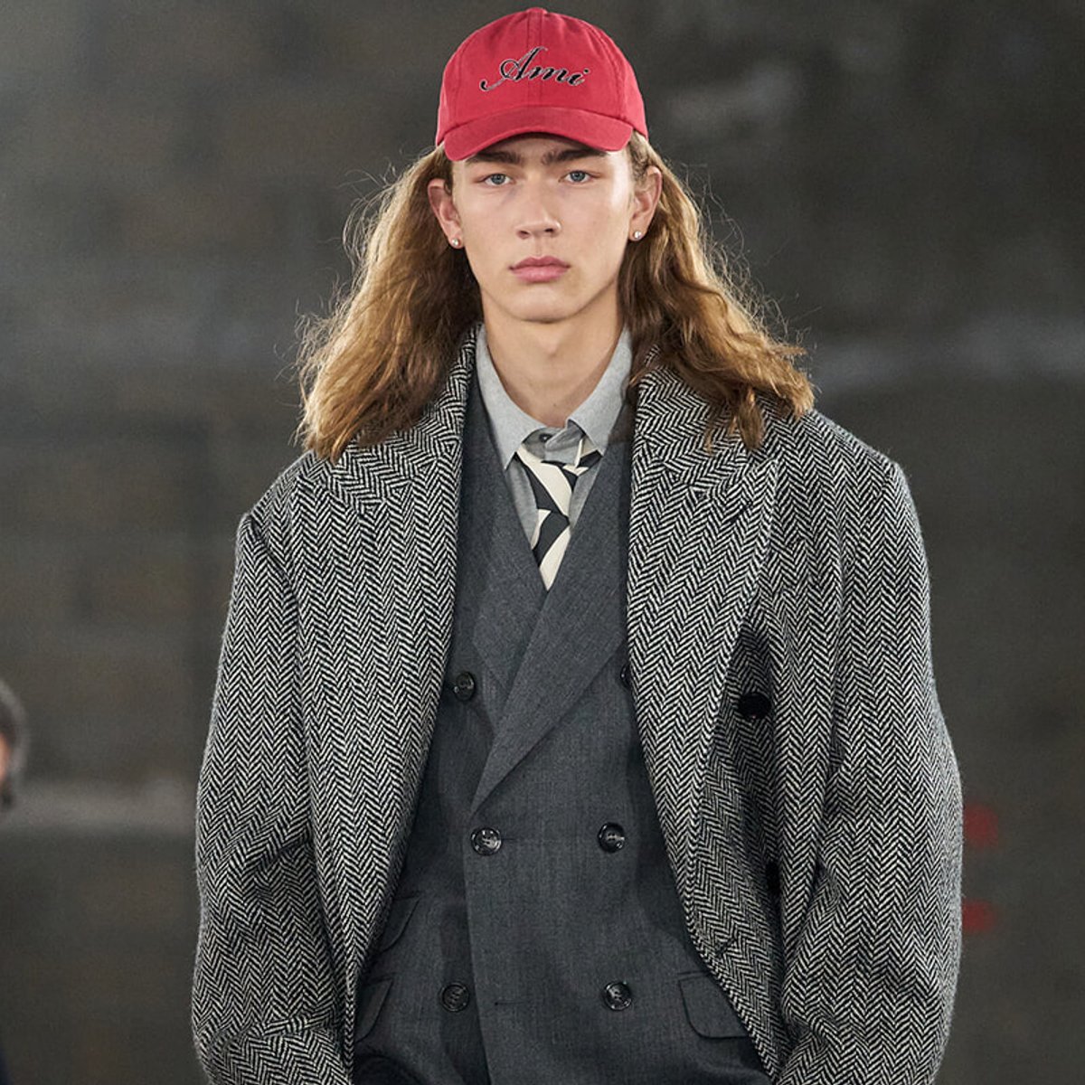 Male model on a runway wearing a gray double‑breasted suit under a textured overcoat and a red cap.