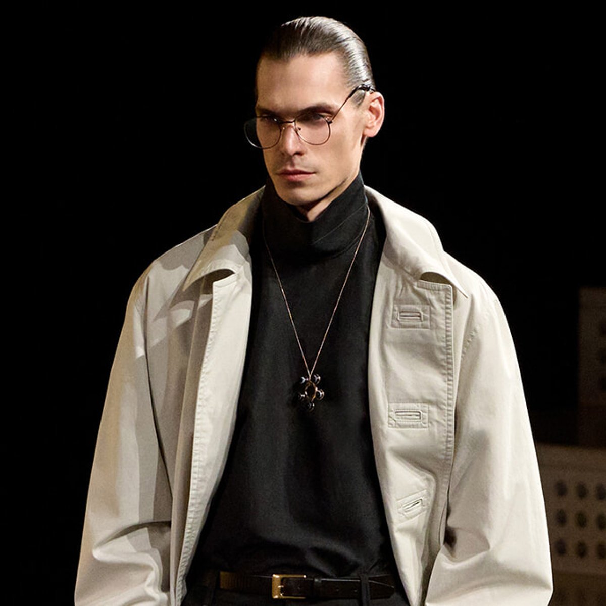Male model with slicked-back hair and round glasses, wearing a beige trench coat over a black turtleneck and a pendant necklace against a dark background.