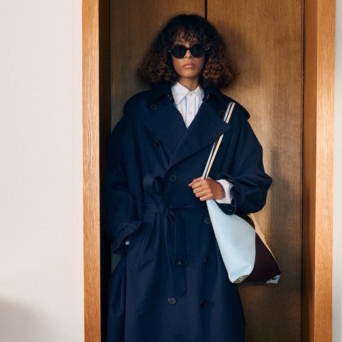 Person wearing a navy trench coat and sunglasses, holding a light blue tote bag, standing in a doorway.