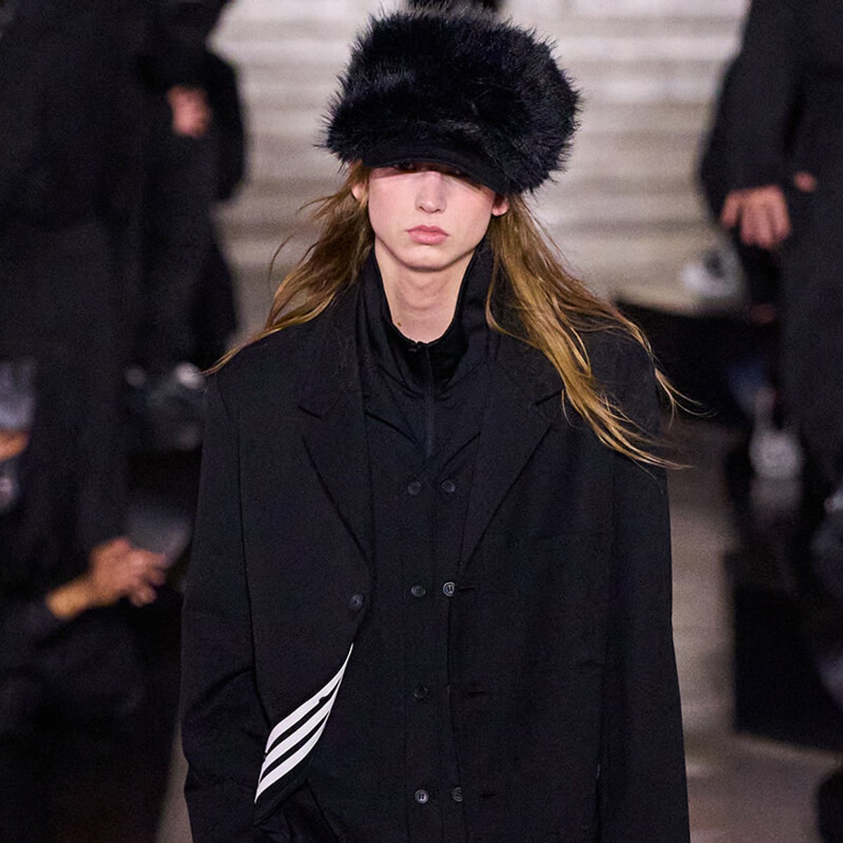 Female model walking a runway in an oversized black coat and a fluffy black fur hat, neutral expression.