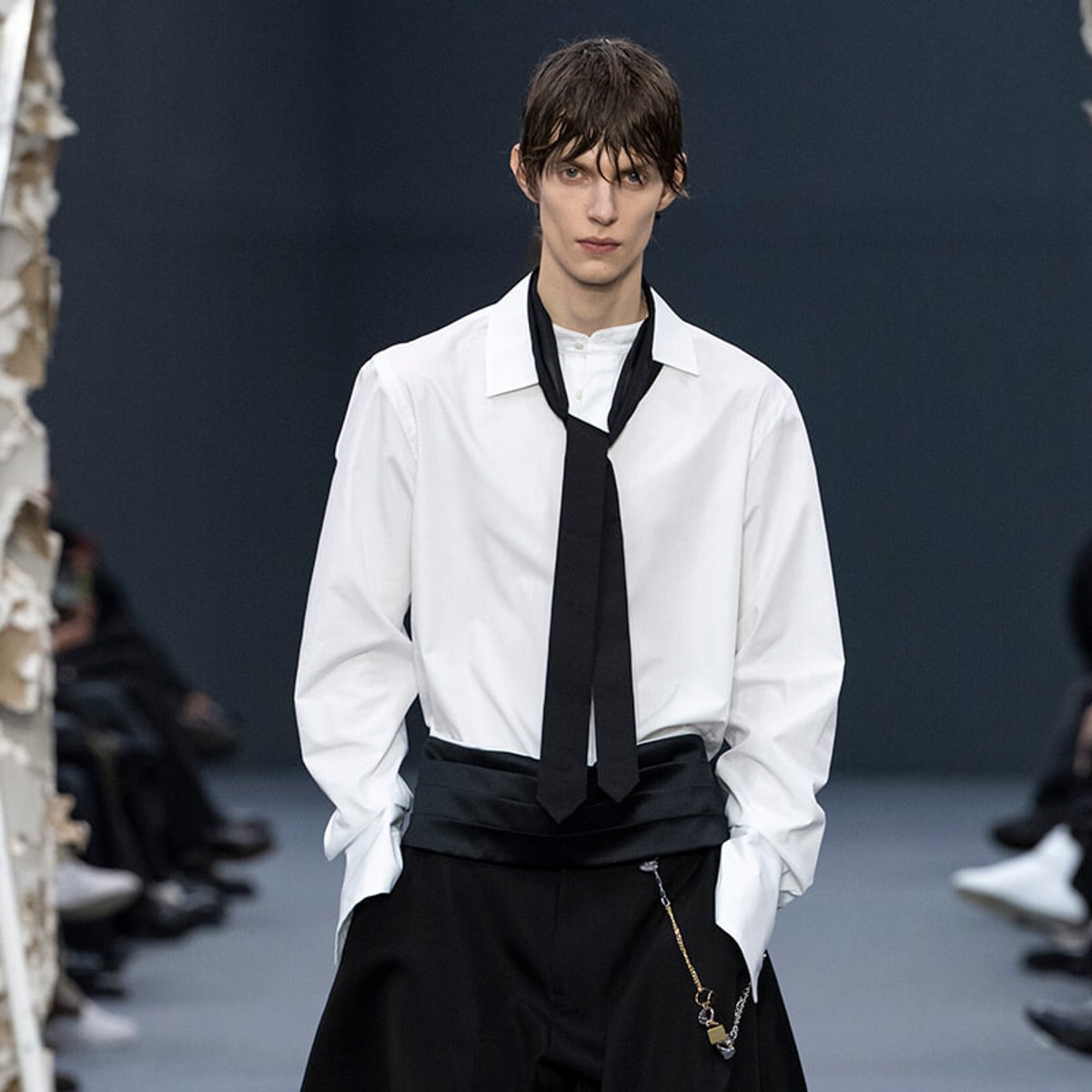 Runway model wearing a white dress shirt with a loose black tie and oversized black pants, with a chain accessory at the hip.