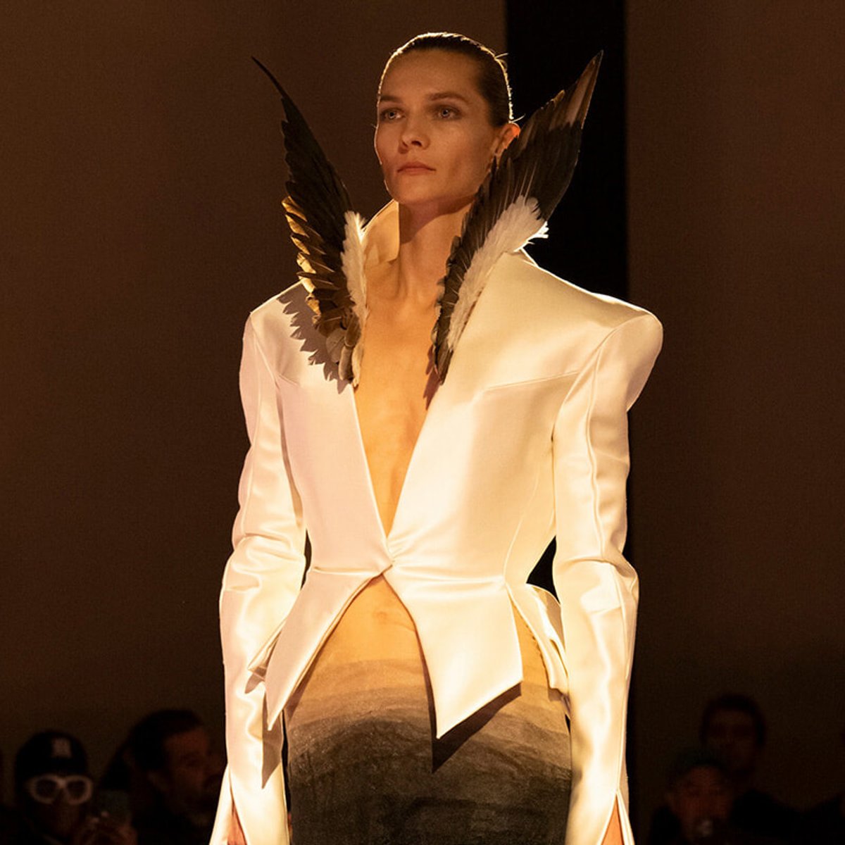 Model walks the runway wearing a white satin blazer with dramatic feathered wing accessories at the shoulders.