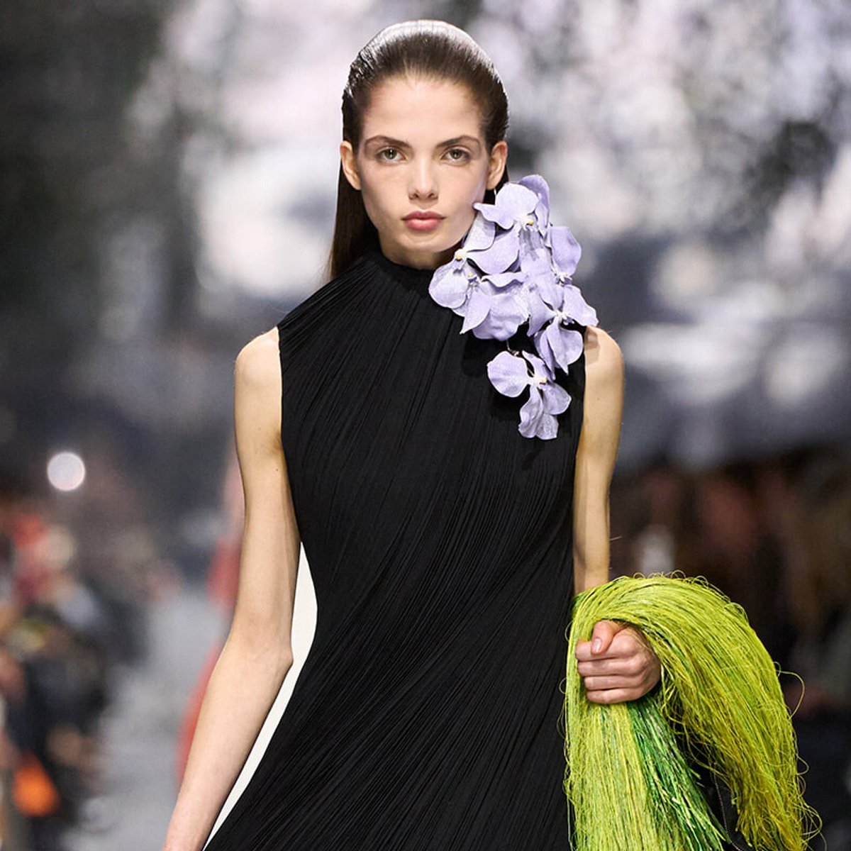 Model walking the runway in a black pleated sleeveless gown with a lavender orchid cluster on the shoulder, holding a bright green fringed accessory.