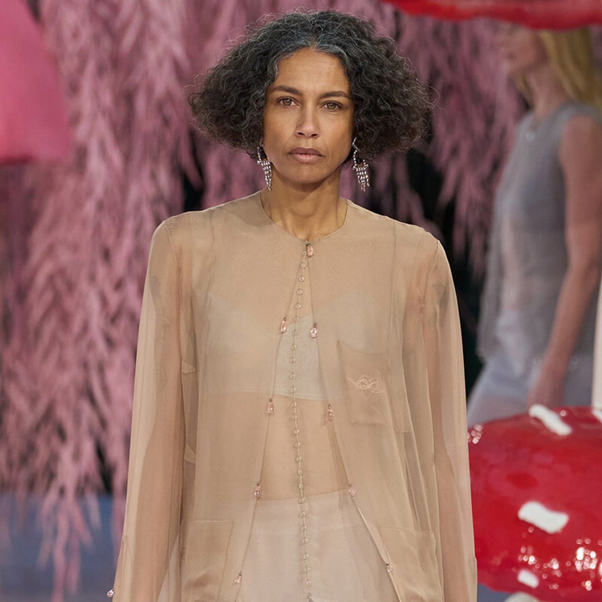 Model on a runway wearing a sheer beige blouse with pink earrings, neutral expression, pink background behind.