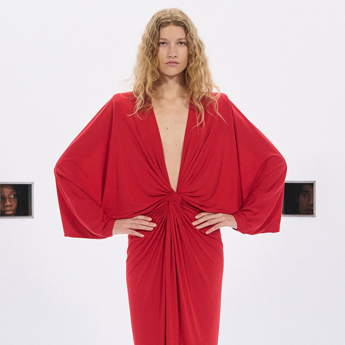 Model in a red draped gown with voluminous sleeves, standing with hands on hips against a white backdrop.